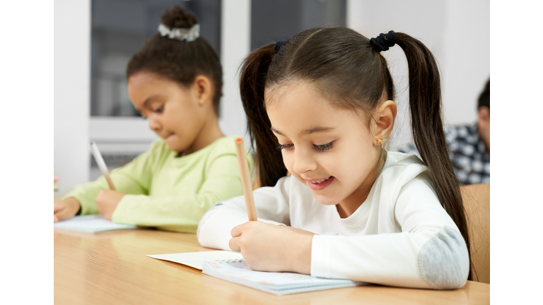 Side view of attractive happy student sitting at table and writing in copybook. Small female student keeping pen and drawing, learning and studying at school. Concept of knowledge.