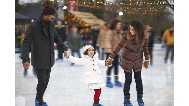 Girl holding parents hands ice skating, smiling