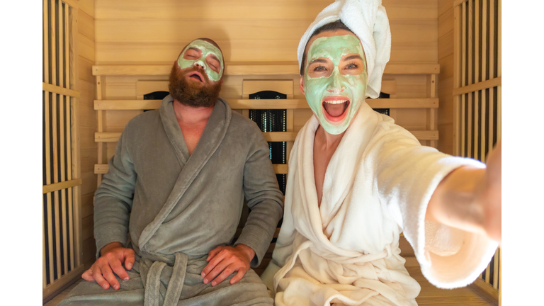 A young funny couple having relax SPA day in sauna, a woman making selfie with beauty mask while her boyfriend is asleep.