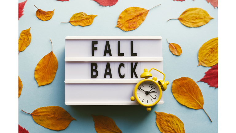 Yellow retro alarm clock, autumn red and orange leaves over blue background. Fall back, daylight saving time concept.