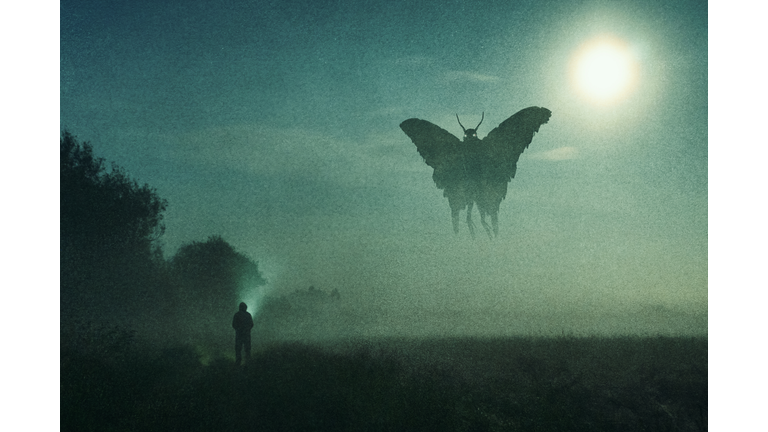A horror concept. Of a man looking at a mysterious monster Mothman figure, flying in the sky. Silhouetted against the moon at night. With a grunge, textured edit.