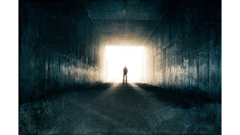 A silhouette of a blurred figure emerging from the light at the end of a dark sinister tunnel. With a grunge, vintage, grainy edit.