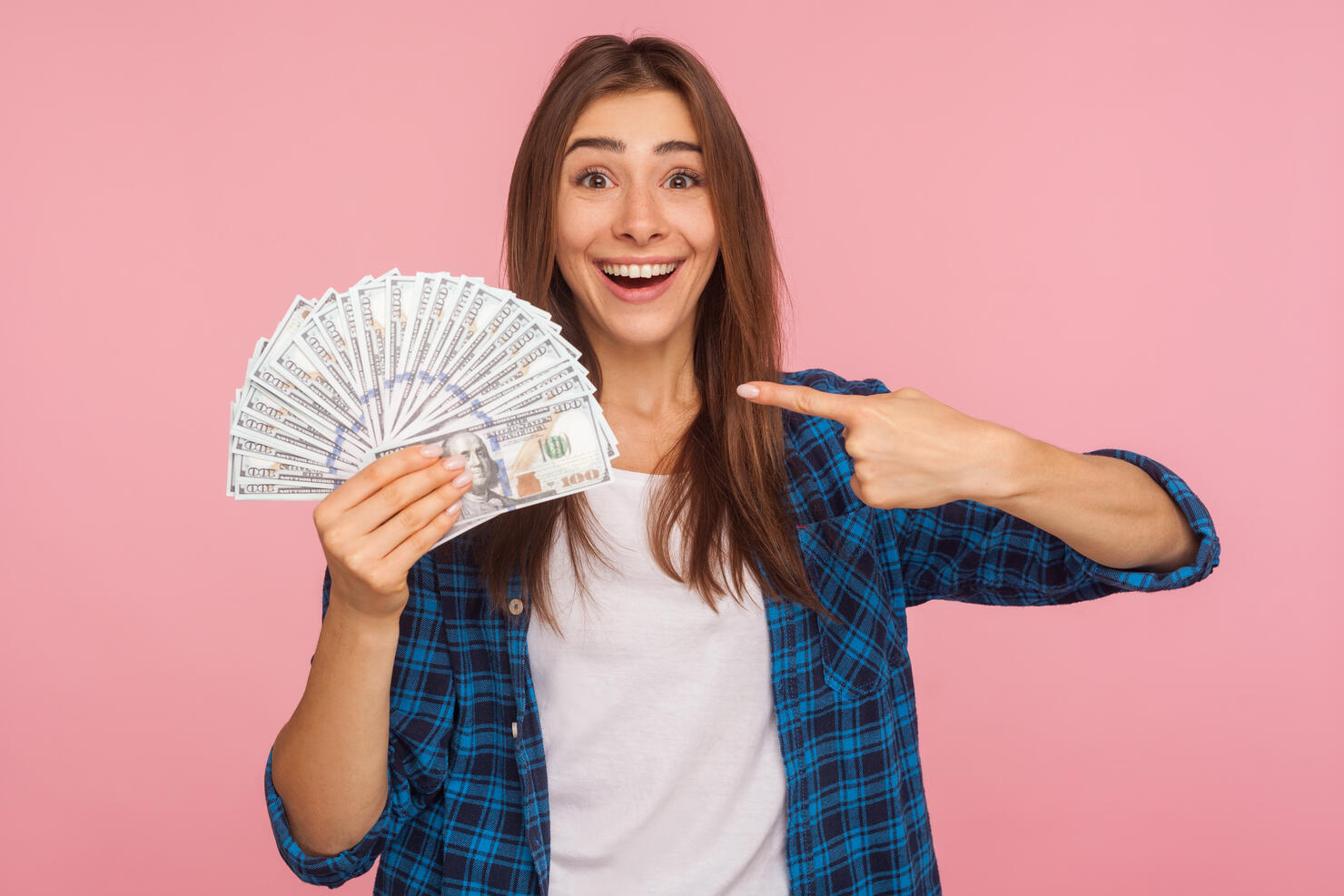 Portrait of joyful lucky girl in casual shirt happy to hold lot of money, pointing at dollar bills in hand and smiling
