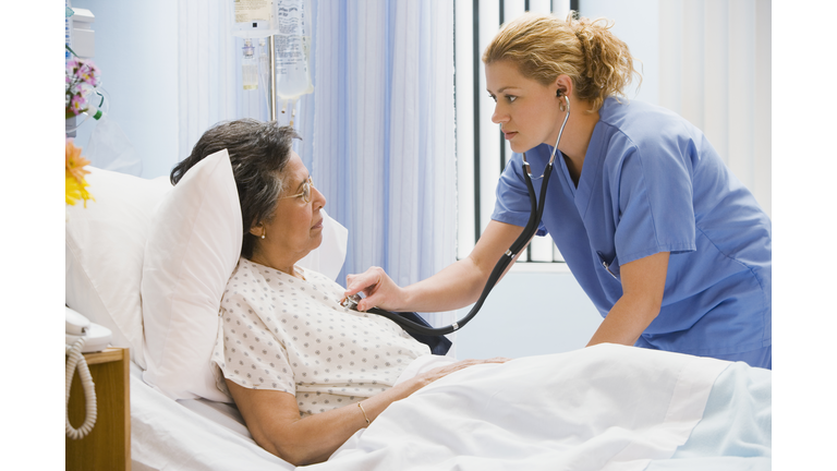 Female nurse using stethoscope on senior woman in hospital bed