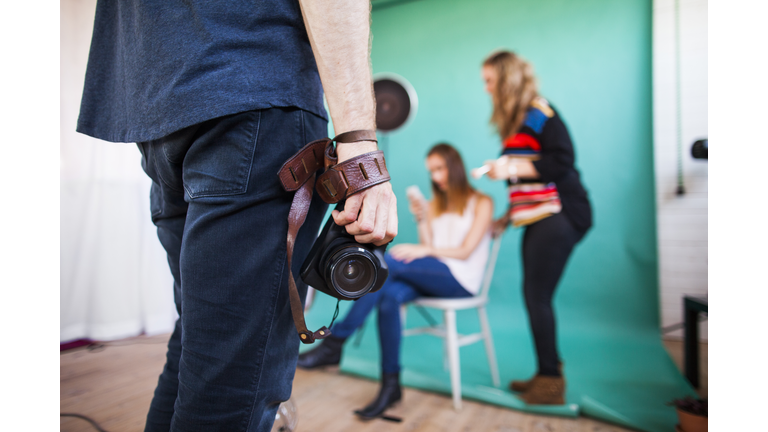 Midsection of photographer holding digital camera while models getting dressed in background at studio