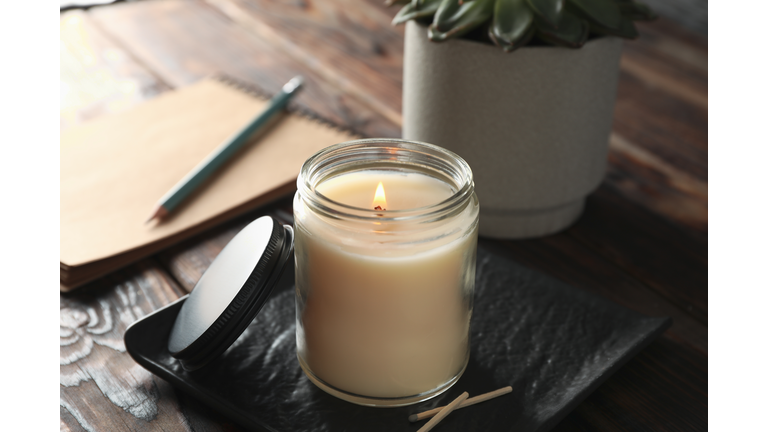 Burning candle in glass jar, succulent and notebook on wooden background, close up