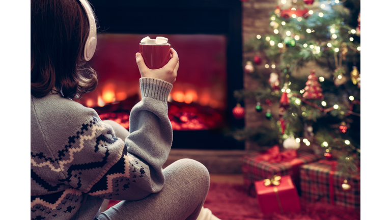 Woman in headphones with cup of hot cocoa and marshmallow sitting and warming at winter evening near fireplace flame and  christmas tree.