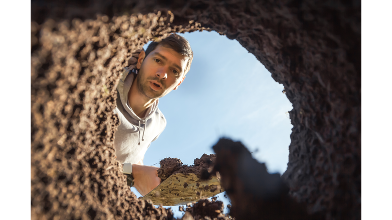 A young guy digging a deep hole with a shovel is surprised by an amazing find. View from the underground at the grave digger