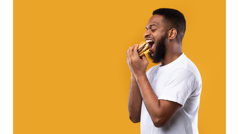 Black Millennial Guy Biting Burger Eating Over Yellow Background, Side-View