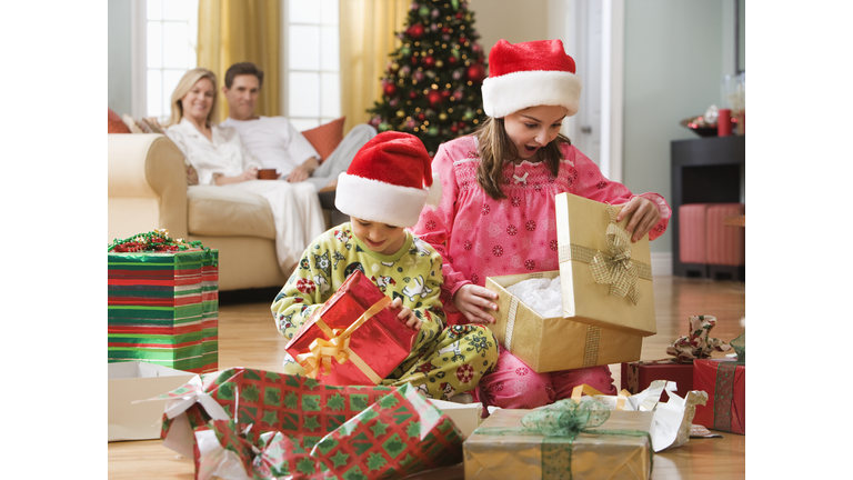 Boy (6-7 years) and girl (8-9 years) unwrapping Christmas presents in living room, parent sitting in background