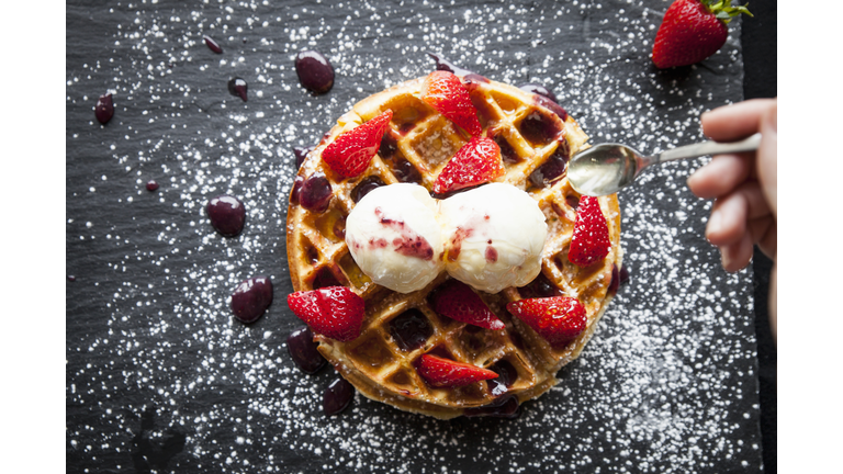 Female hand holding spoon over strawberries and ice cream waffle on slate