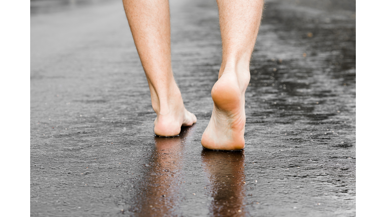 Young man's barefoot walking on the wet, dark black asphalt after warm rain. Cloudy day in summer. Back view.
