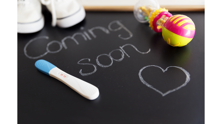 Waiting for labour, maternity concept. "Coming soon" words on black chalkboard with positive pregnancy test, white baby shoes and rattle