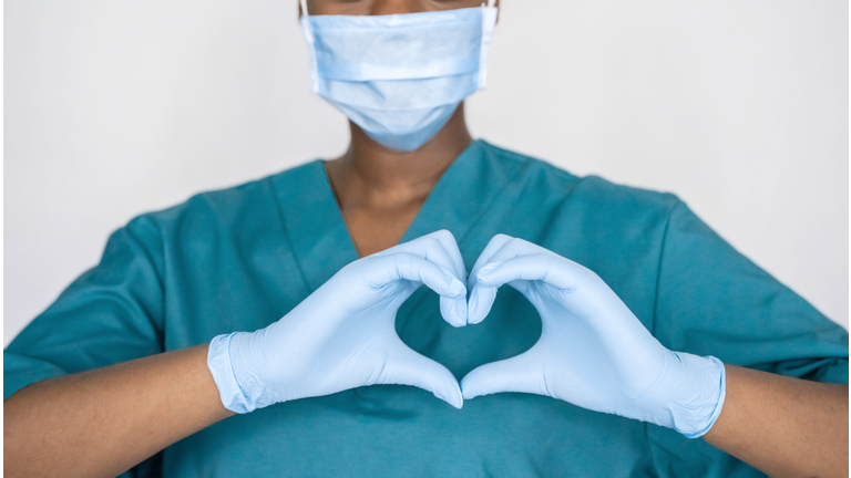 Female african professional medic nurse wear face mask, gloves, blue green uniform showing heart hands shape. Medical love, care and safety symbol, corona virus health protection sign concept. Closeup