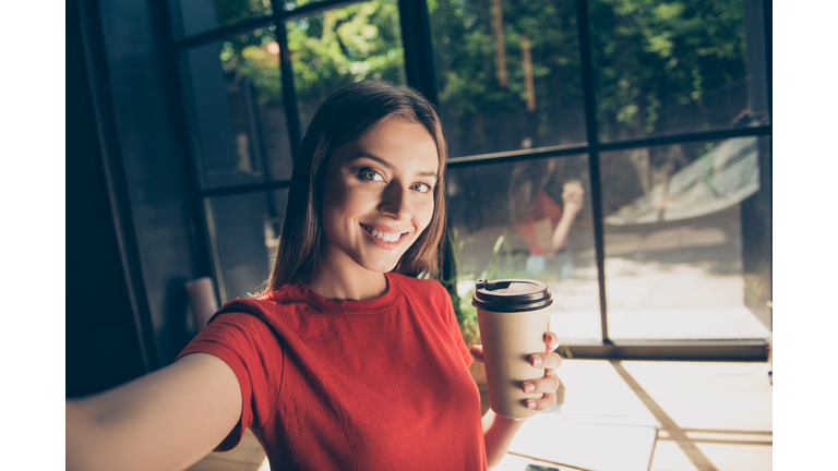 Beautiful young woman doing selfie on the front camera of her smartphone and holding a paper cup with a drink