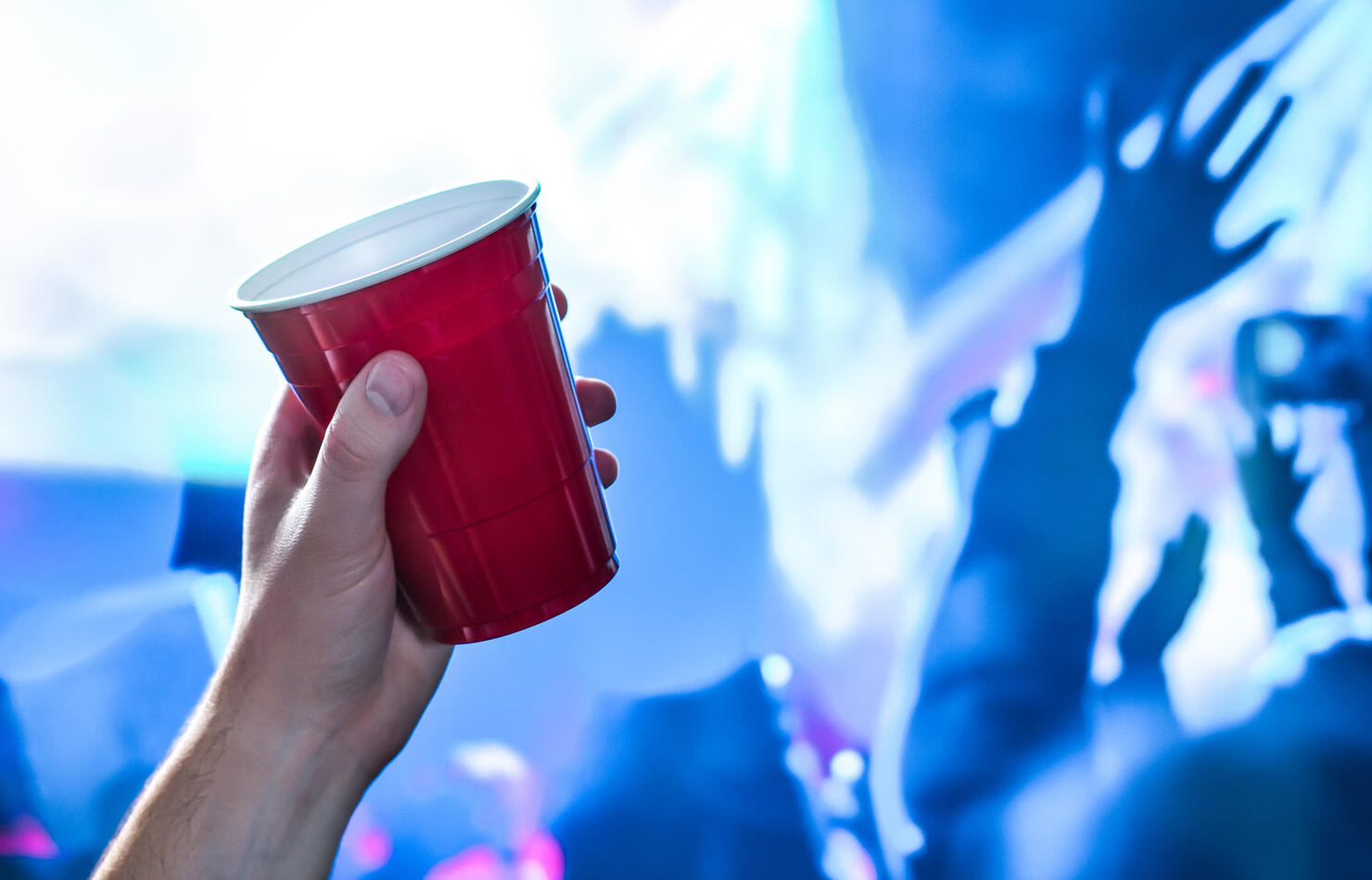 Red party cup in hand in night club, bar or college student event. Plastic beer mug. People having fun in blue nightclub disco lights on dance floor