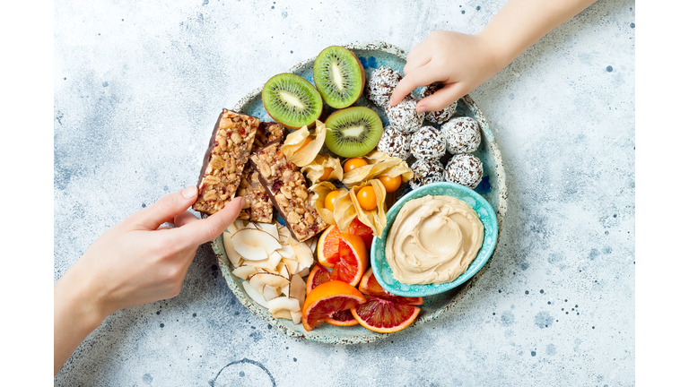 Mother giving healthy vegan dessert snacks to toddler child. Concept of healthy sweets for children. Protein granola bars, homemade raw energy balls, cashew butter, toasted coconut chips, fruits platter