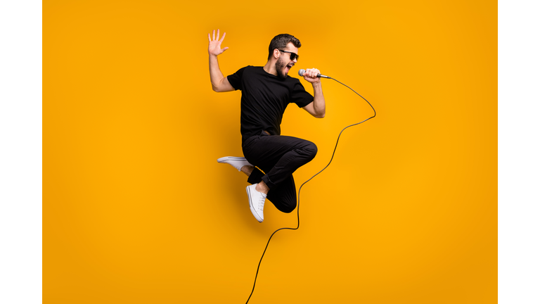 Full body profile photo of crazy hipster guy jumping high holding microphone music lover singing favorite song wear sun specs black t-shirt pants isolated yellow color background
