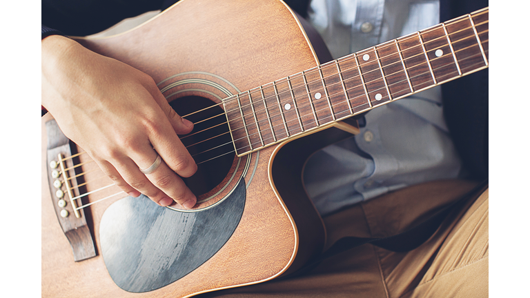 Stylish, fashionable man in a blue shirt, dark blue jacket and brown pants playing guitar. The concepts of hobby, passion and interest in music. Hands guy touch the strings of the guitar, close-up.