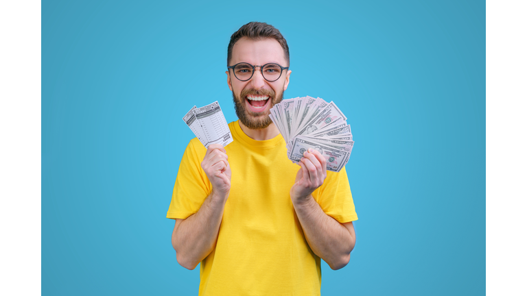 Portrait of happy euphoric bearded man in yellow t-shirt posing with lottery ticket and ward of dollar bancknotes in hands as betting, gambling, money win concept