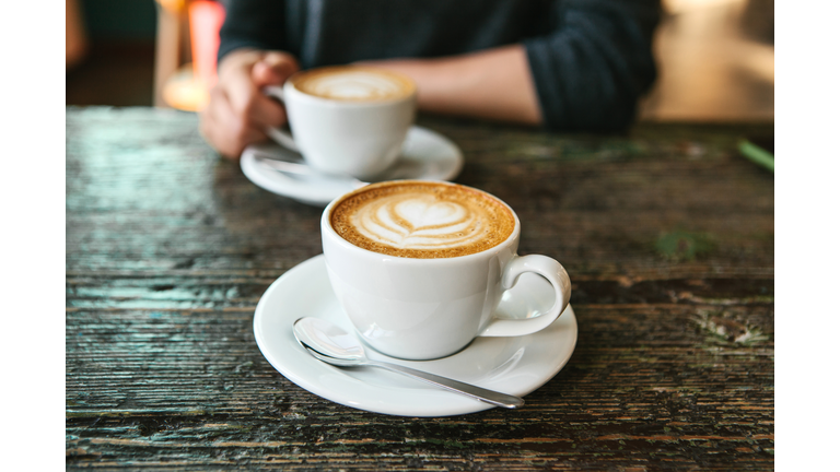 Two cups of coffee on a wooden table, the girl holds in her hand one cup of coffee in the background. A photo indicates a meeting of people and a joint pastime.