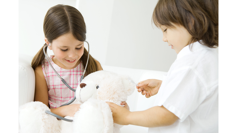 Two children playing doctor, girl listening to teddy bear's heart with stethoscope