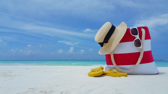 Tourism and travel, tropical vacation concept, striped beach bag flip-flops glasses and children toy on white sand beach. Family vacation background