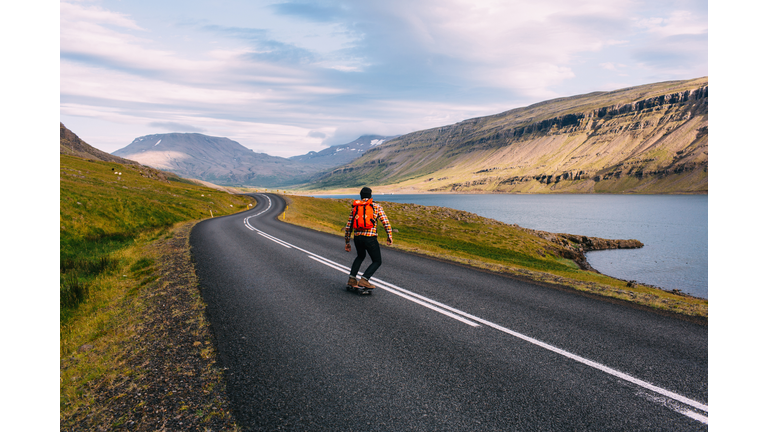Rear view of mid adult man skateboarding on curving open road by lake, Iceland