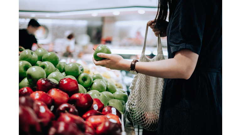 Cropped shot of young Asian woman shopping for fresh organic groceries in supermarket. She is shopping with a cotton mesh eco bag and carries a variety of fruits and vegetables. Zero waste concept