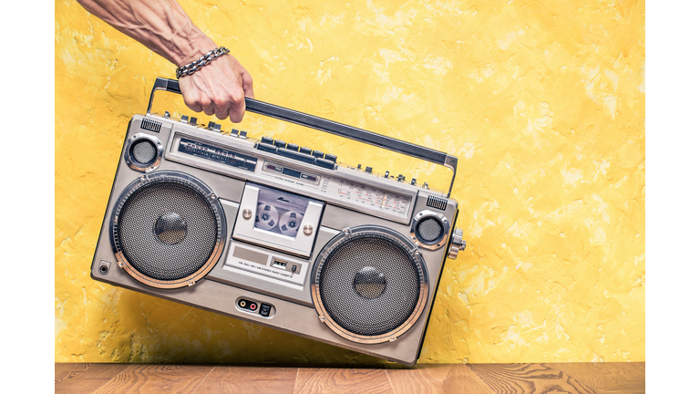 Retro outdated portable stereo boombox radio receiver with cassette recorder from circa 1980s in a strong man's hand front concrete textured yellow wall background. Vintage old style filtered photo