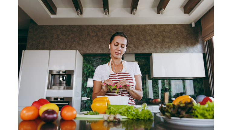 Young woman with headphones in the ears holding vegetables in hands in kitchen with laptop on the table. Vegetable salad. Dieting concept. Healthy lifestyle. Cooking at home. Prepare food