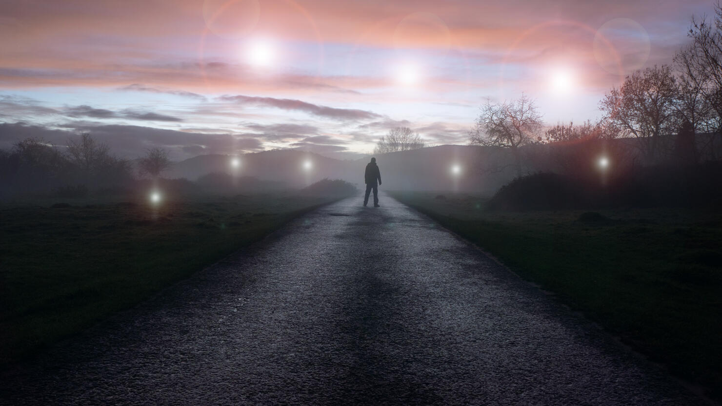 A UFO concept of glowing orbs, floating above a misty winters road just after sunset. With a silhouetted figure , back to camera, looking at the lights.