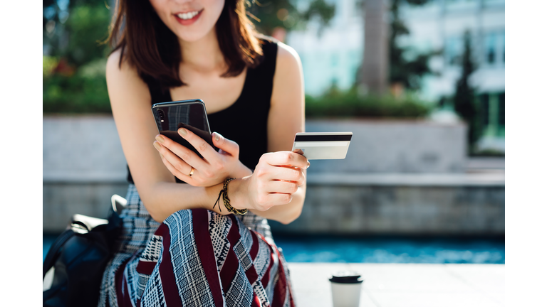 Beautiful smiling young Asian woman sitting in an urban park, enjoying coffee, shopping online with smartphone and making mobile payment with credit card on hand. Online shopping and mobile payment concept