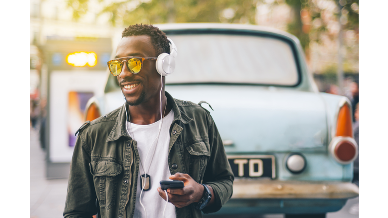 Smiling young man wearing sunglasses listening music through headphones against vintage car in city