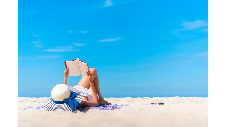 Woman Lying While Reading Book On Beach Against Blue Sky