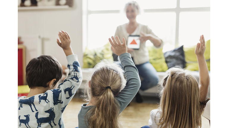 Rear view of students raising hands while teacher showing digital tablet at preschool