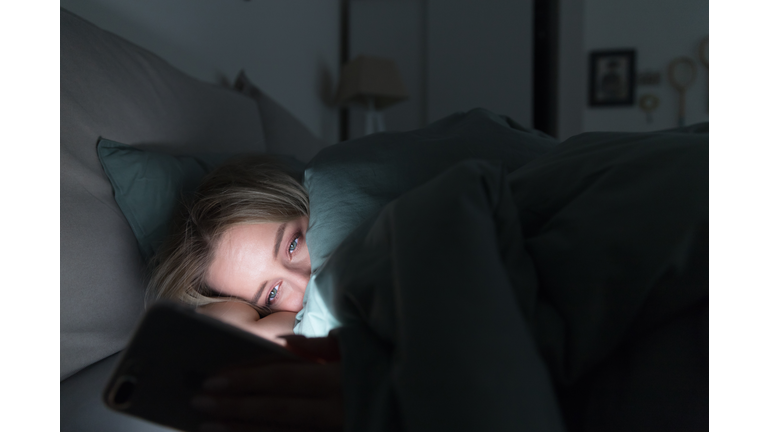 Sleepy Tired Woman Lying In Bed Under The Blanket Using Mobile Phone At Night. Insomnia. Addiction.