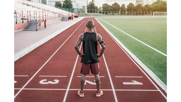 Athlete standing at the start line with hands on waist. Rear view of runner standing on running track.