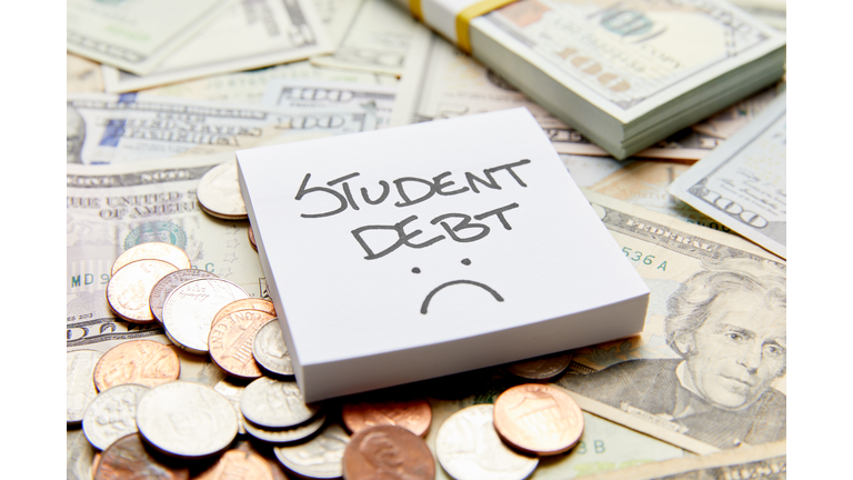 Student debt with sad face written on white sticky note on top of cash money with stack of money and coins