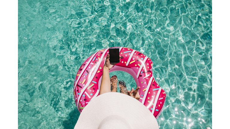 top view of a woman relaxing in the pool with pink donuts in hot sunny day. Summer holiday idyllic. Enjoying suntan Woman in bikini and a hat. Holidays and summer lifestyle. She is using mobile phone