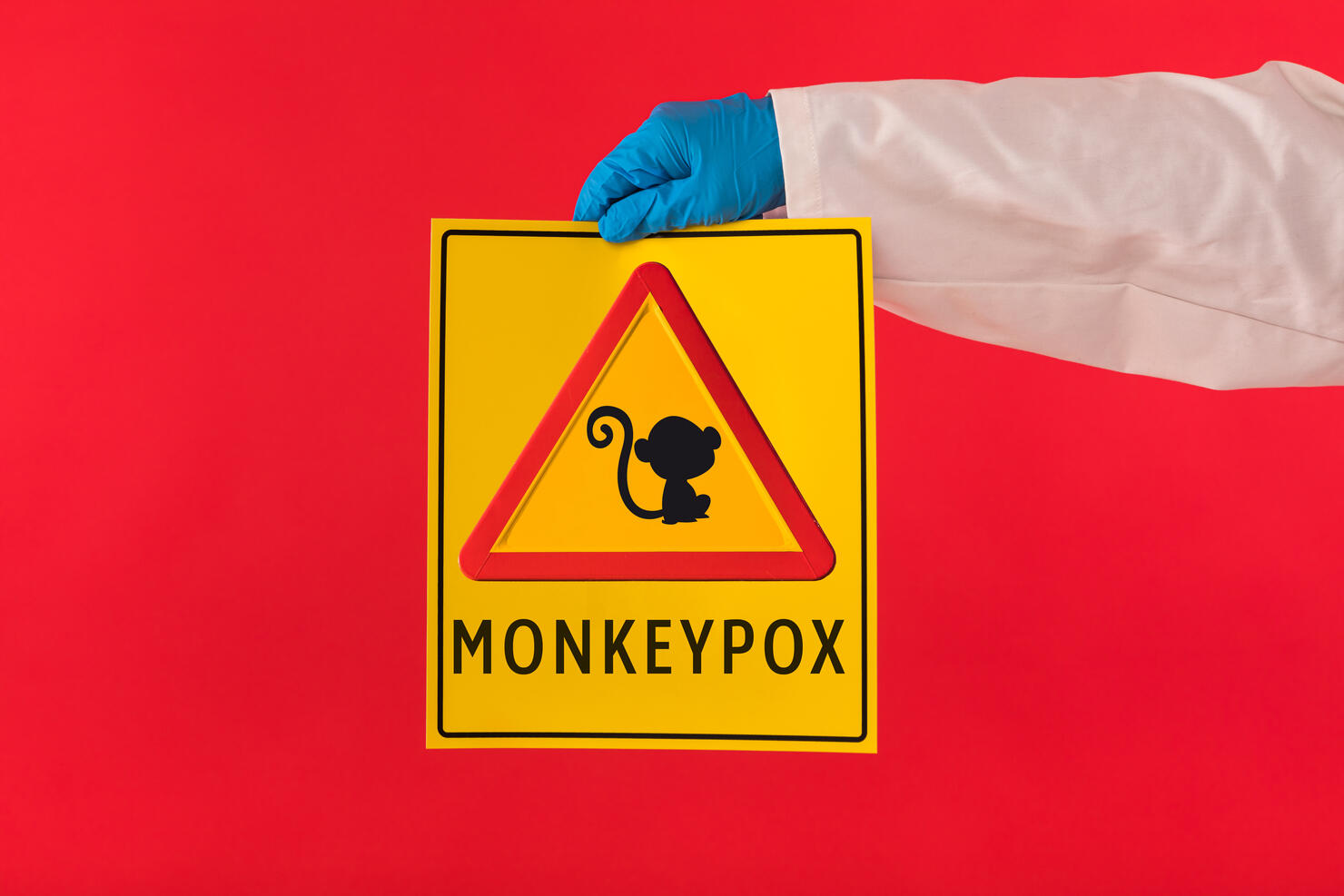 Arm of a doctor wearing a gown and latex gloves holding a yellow sign with a symbol with a monkey danger triangle and Monkeypox, on a red background. Pandemic, virus, epidemic, Nigeria and smallpox.