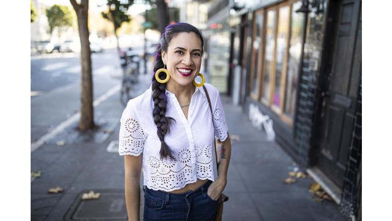 Millennial Latina Stands Alone on City Sidewalk, Smiling and Looking at Camera, Wearing White Lace Blouse and Bright Yellow Hoop Earrings