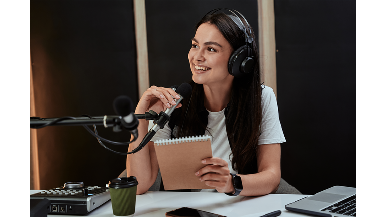 Portait of attractive young female radio host smiling aside while speaking in microphone, moderating a live show