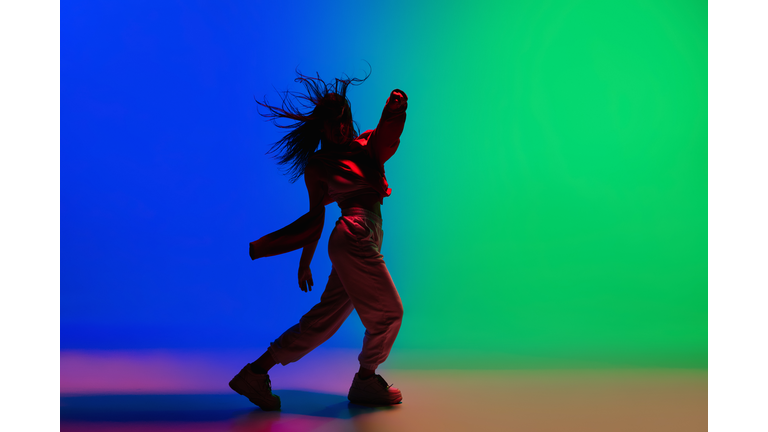 Stylish sportive girl dancing hip-hop in stylish clothes on colorful background at dance hall in neon light. Youth culture, movement, style and fashion, action.