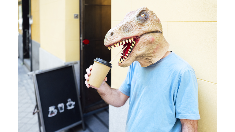 Man wearing dinosaur mask drinking coffee while standing against wall