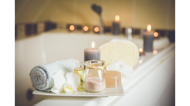 White ceramic tray with home spa supplies in home bathroom for relaxing rituals. Candlelight, salt soap bar, bath salt in jar, massage, bath oil in bottle, blue rolled towel, natural sponge.