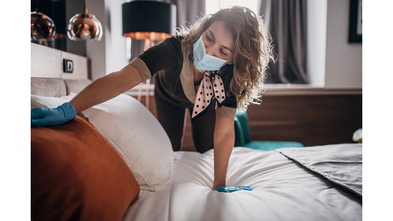 Beautiful maid with medical mask changing bed linen on the bed in a hotel room