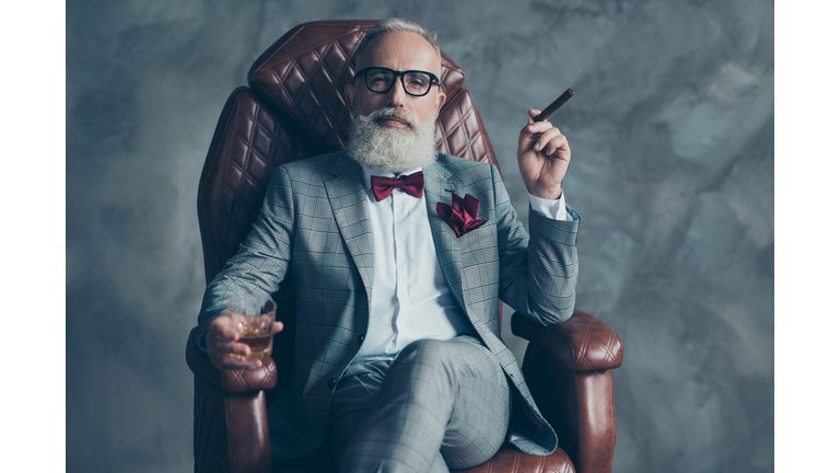 Cool man in glasses, hold cigarette,  glass with brandy, in formal wear, tux with red bowtie and pocket square, sit in leather chair over gray background, looking to the camera, shares, stock, money