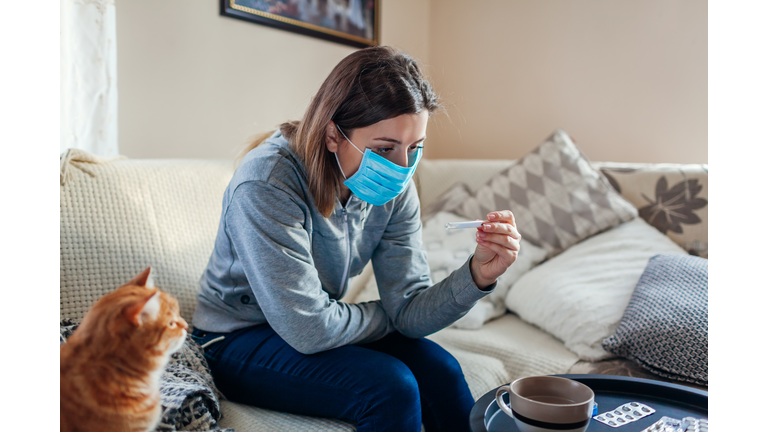 Ill woman checking thermometer having fever. Girl wearing protective mask while having cold, flu at home. Healthcare