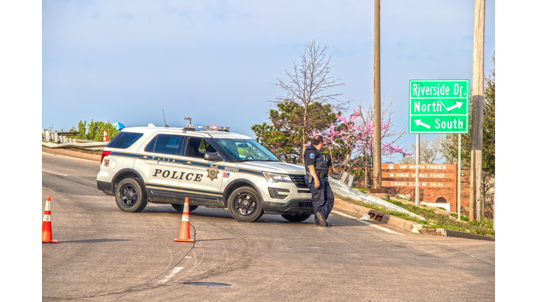 Tulsa cop leans on front of his police car blocking overpass with orange cones on overcast day with damaged railing behind and redwood tree in bloom